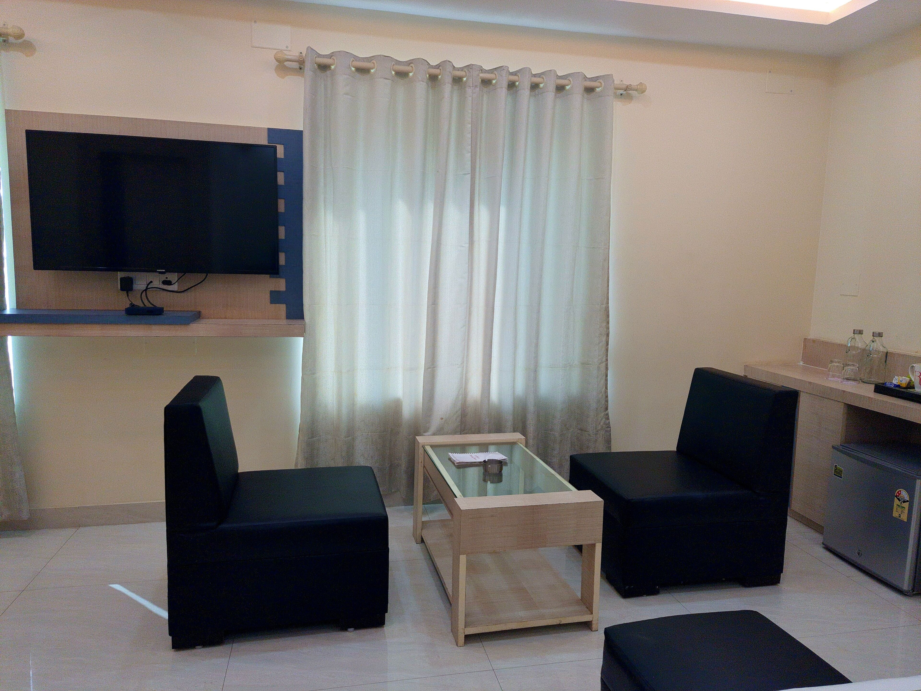 A small, modern living room with two black sofas, a glass coffee table, a flat-screen TV mounted on the wall, and a kitchenette area with a refrigerator and a sink.