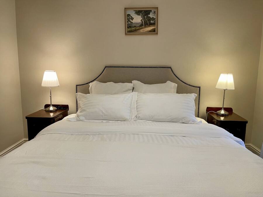 Una cama bien hecha con ropa de cama blanca, dos lámparas de noche y un cuadro de paisaje colgado sobre la cabecera.
