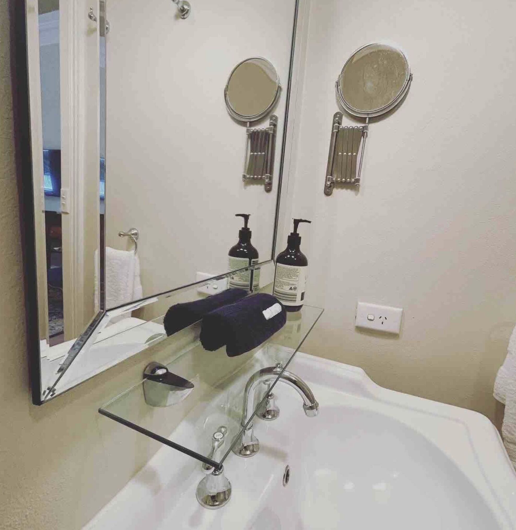 A bathroom with a large mirror, two round mirrors, a white sink, a glass shelf with toiletries, and a towel rack.