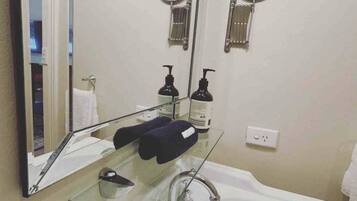 A bathroom with a large mirror, two round mirrors, a white sink, a glass shelf with toiletries, and a towel rack.