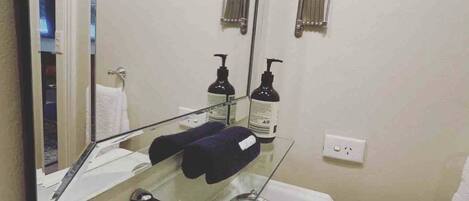 A bathroom with a large mirror, two round mirrors, a white sink, a glass shelf with toiletries, and a towel rack.