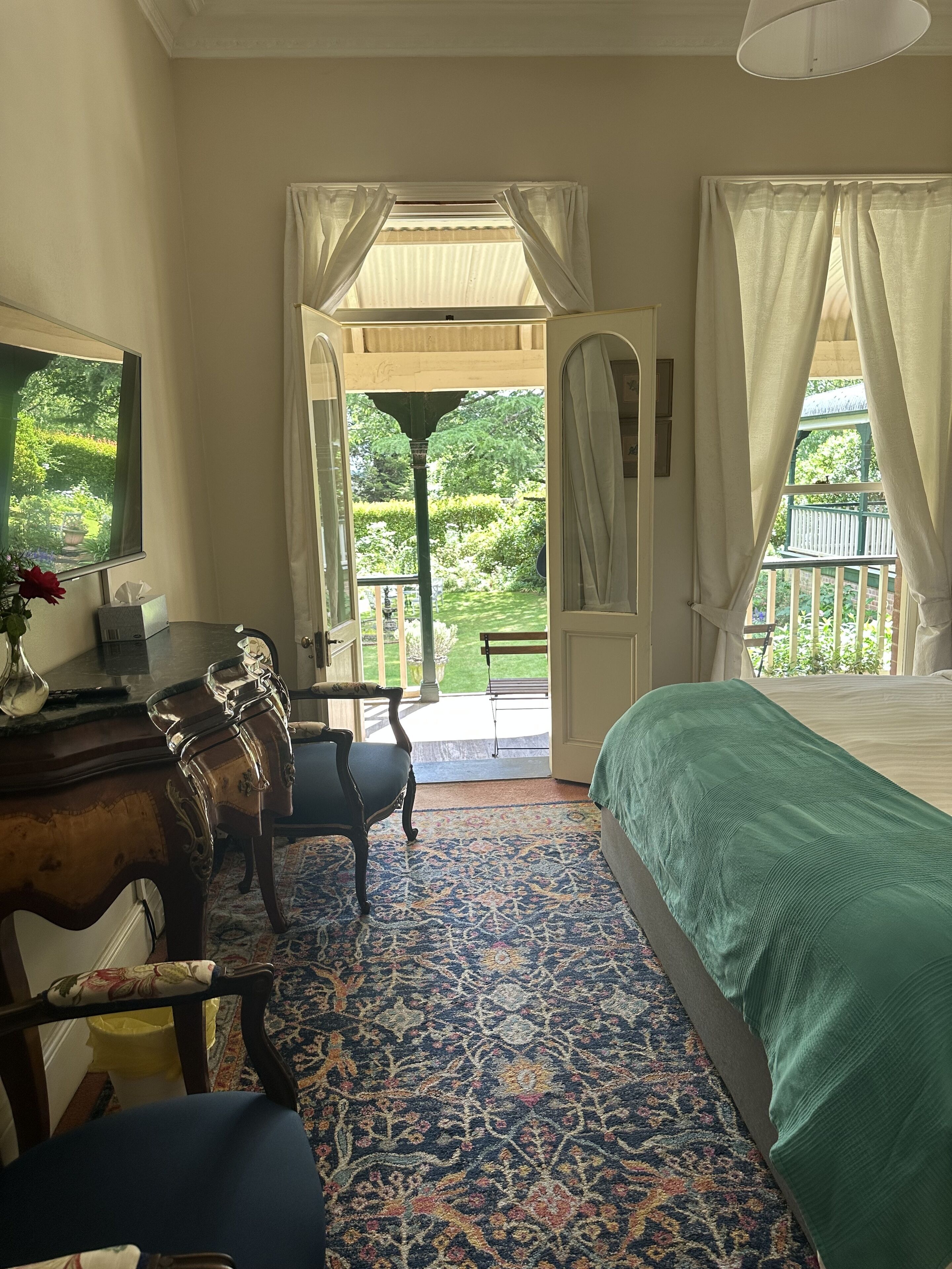Un dormitorio con una cama, un escritorio con silla, un televisor y vista a un jardín por una puerta abierta.