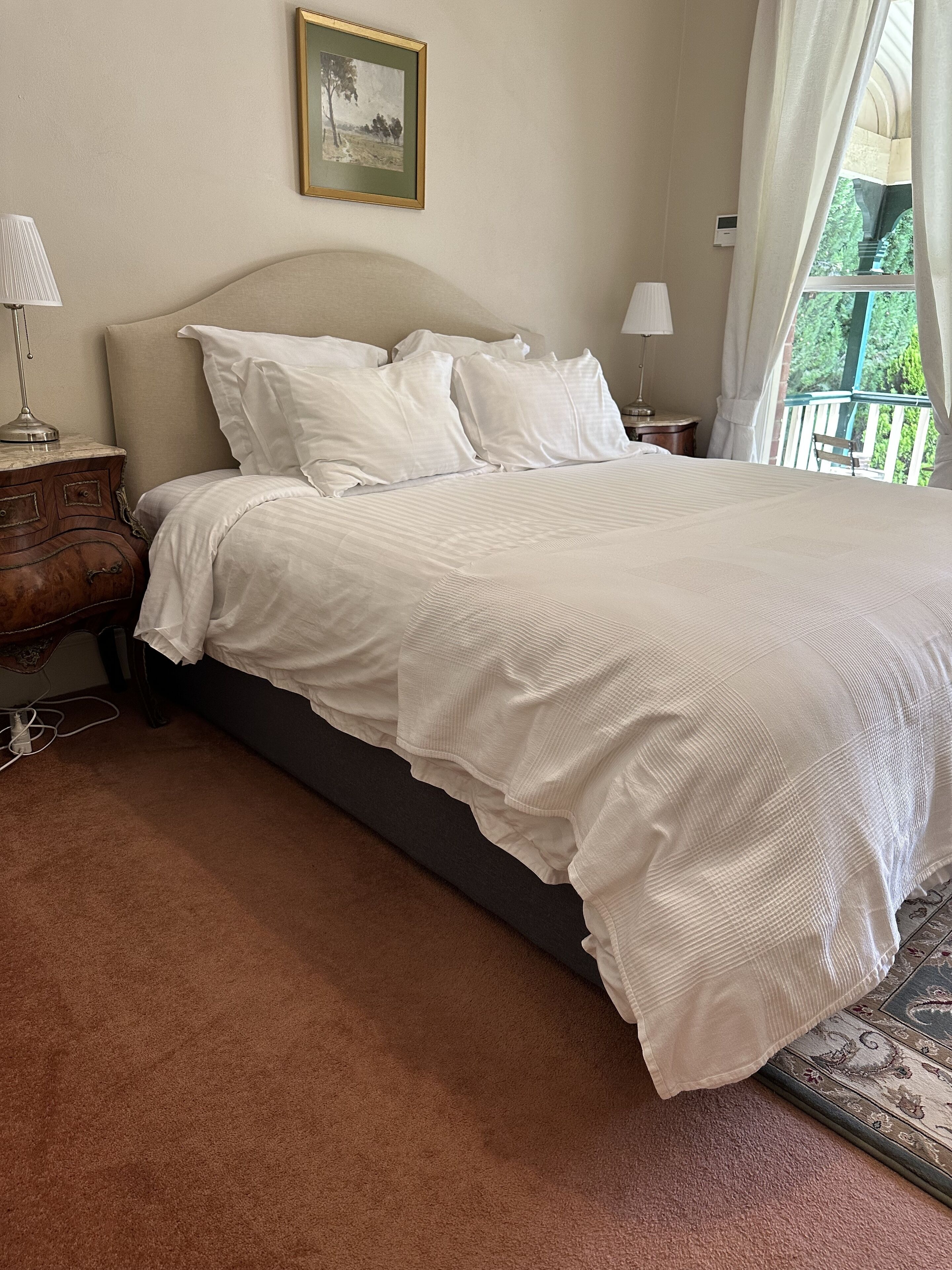 A bedroom with a bed, nightstands, a lamp, a framed picture on the wall, and a view of greenery outside.