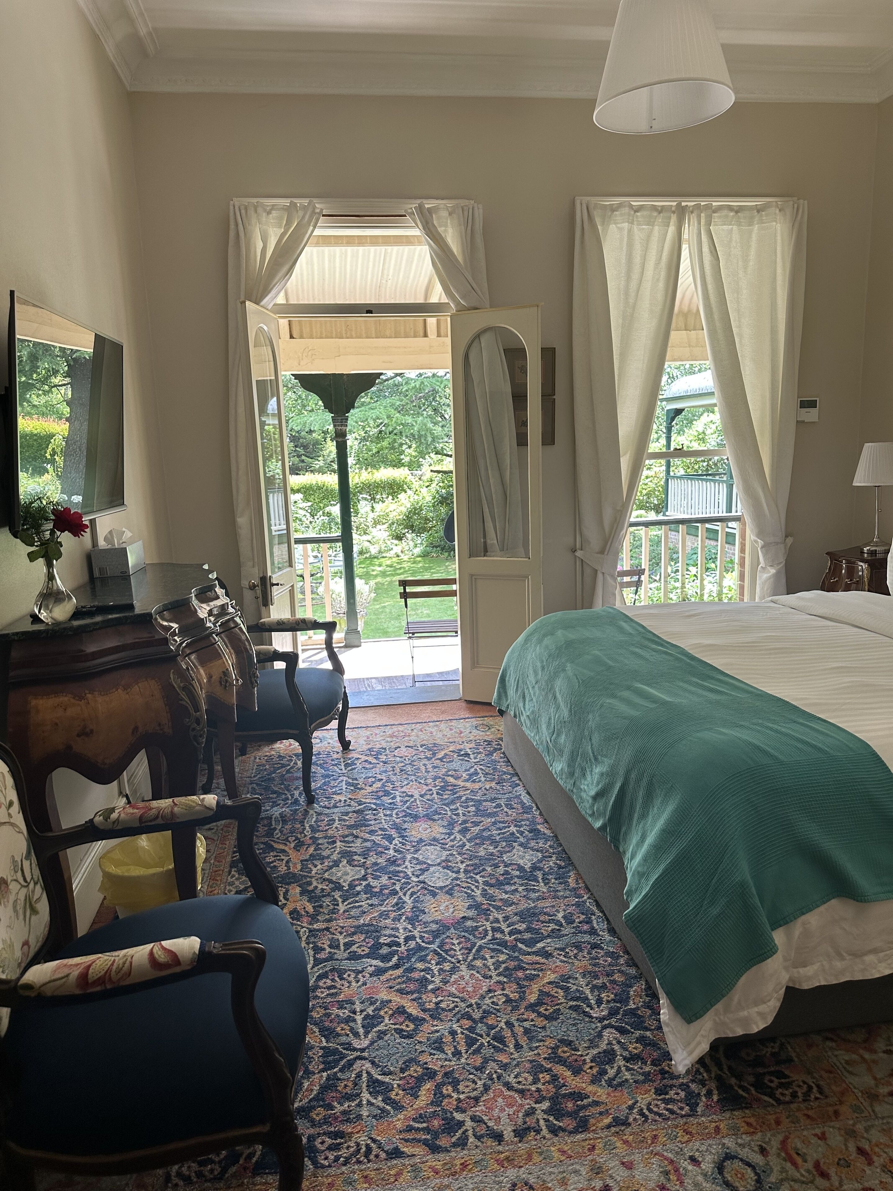 A bedroom with a large bed, a desk with a chair, a small table with a vase, and a view of a garden through a door.