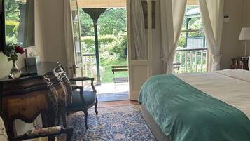 A bedroom with a large bed, a desk with a chair, a small table with a vase, and a view of a garden through a door.