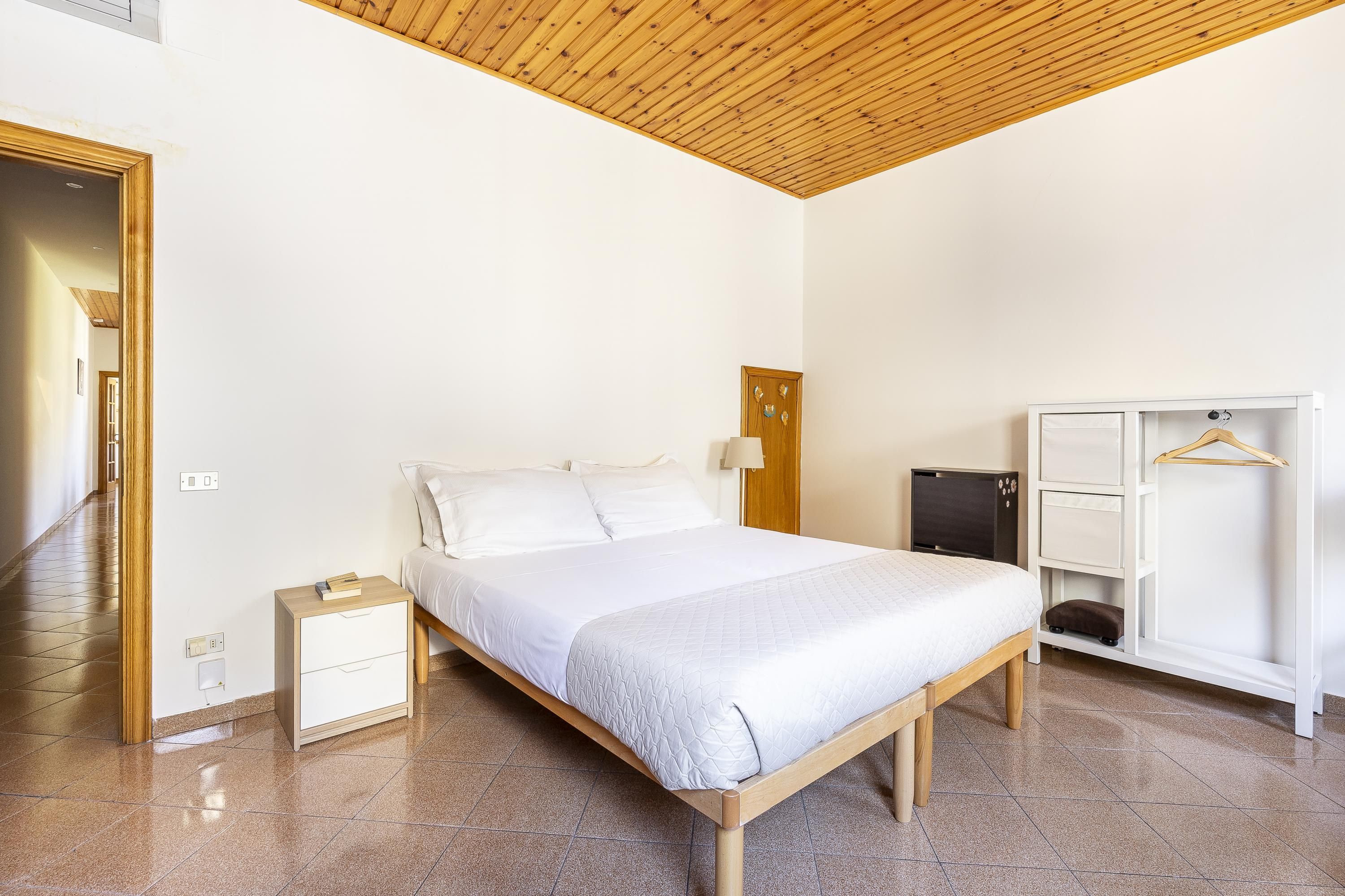 Un dormitorio con un cabecero de madera, una mesita de noche blanca, un armario y una puerta que da a otra habitación.