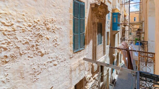 A narrow street with buildings featuring traditional architecture, including stone walls and wooden shutters. - Central Valletta Townhouse (Valletta)