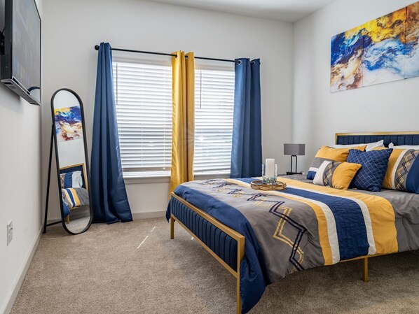 A bedroom with a bed, a mirror, a window with curtains, a painting on the wall, and a lamp on a side table.
