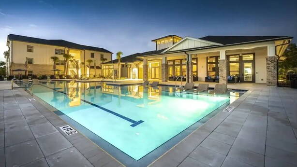 Ein Swimmingpool mit Fliesen umgeben, ein überdachter Sitzbereich und ein Gebäude mit großen Fenstern und einer flachen Dachkonstruktion.