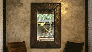 A rustic room with a concrete wall, a framed picture, and two wooden chairs around a small table with a yellow flower. - Tamuku Padang Padang, CARI Surf Camp (Pecatu)
