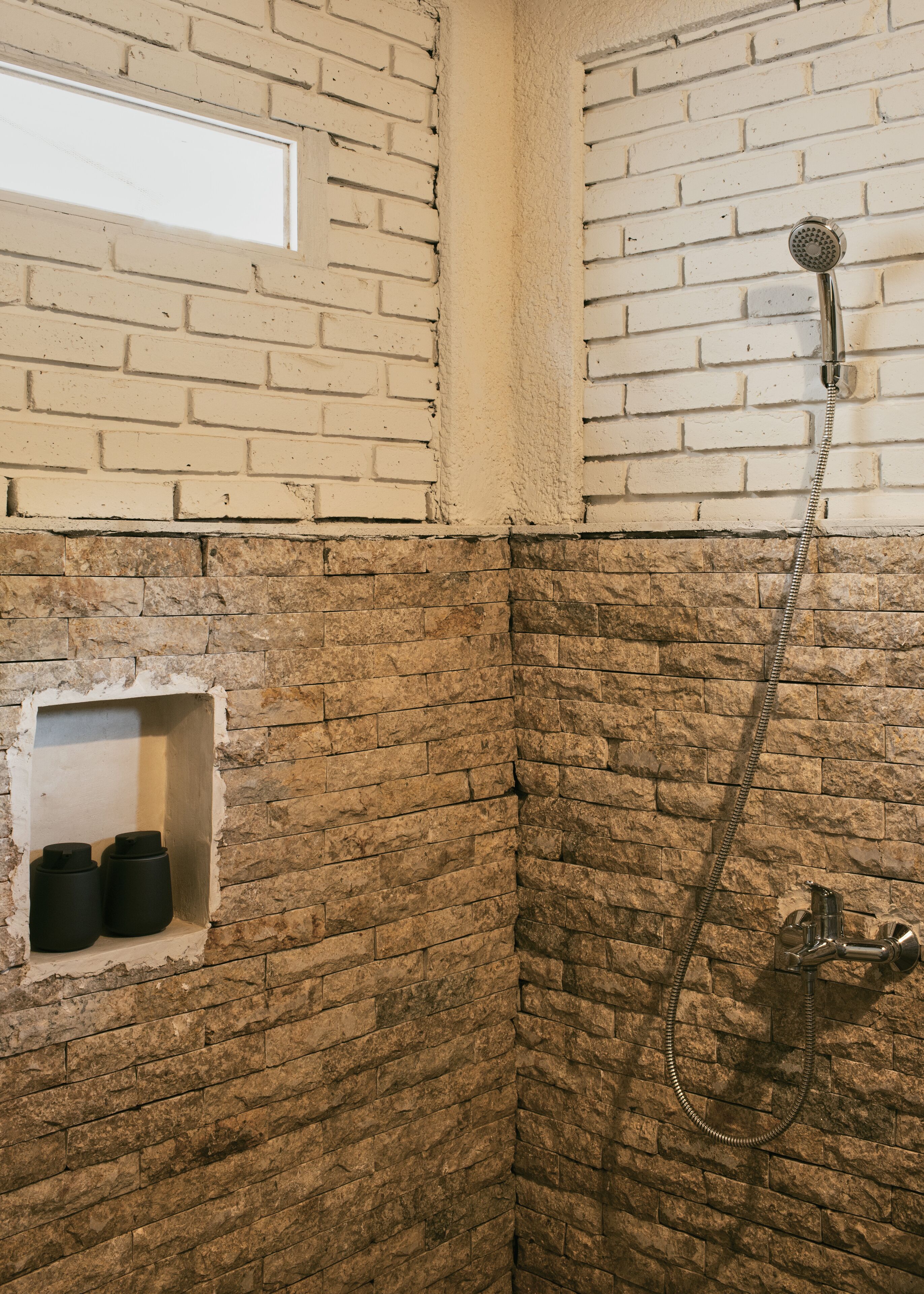 A shower area with a brick wall, a showerhead, and two black containers on a shelf.