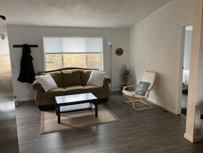 A living room with a beige sofa, a wooden coffee table, a wooden chair, a potted plant, and a window with blinds.