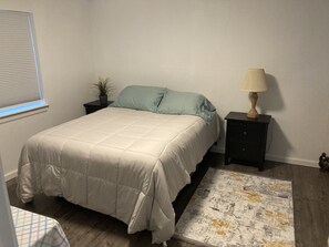 A bedroom with a bed, a nightstand, a lamp, a window with blinds, and a small plant.