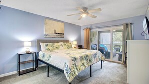 Une chambre à coucher comprenant un lit, un ventilateur de plafond, une table de chevet, une lampe, un tableau et une porte-fenêtre coulissante donnant sur un balcon.