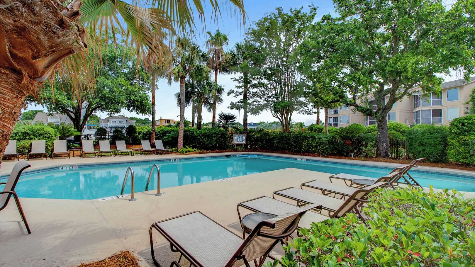 A swimming pool with lounge chairs, palm trees, and a building in the background.