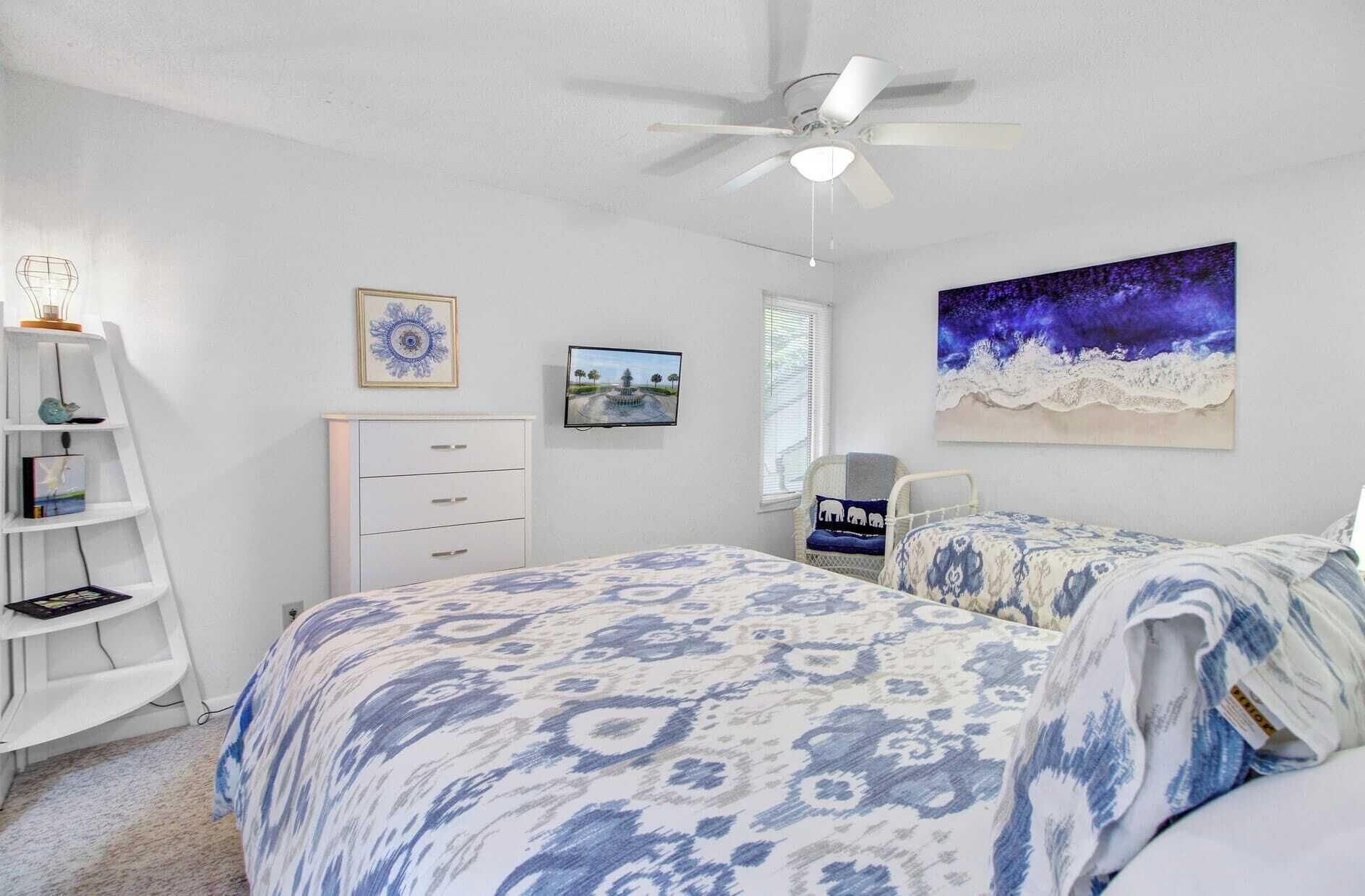 Une chambre à coucher comprenant un lit, une commode, une chaise, un ventilateur de plafond et un mur orné d’un tableau.