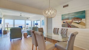 A spacious dining area with a glass table and cushioned chairs, a chandelier, and a painting of boats on the wall.