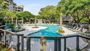 A swimming pool with lounge chairs and palm trees, surrounded by a fence and buildings.