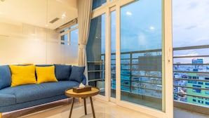 Un salón moderno con un sofá azul, cojines amarillos, una pequeña mesita de madera y un amplio ventanal con vistas a la ciudad.
