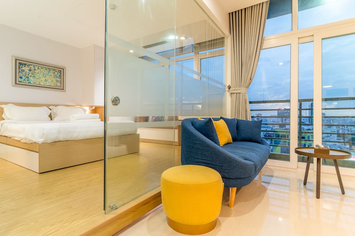 A modern hotel room with a glass partition, a bed, a sofa, and a view of the city.