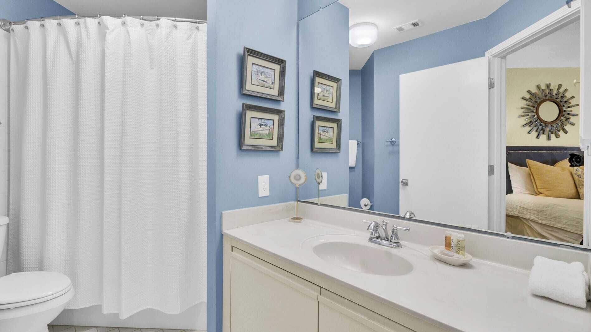 A bathroom with a white toilet, a white sink, and a shower curtain. There are three framed pictures on the wall and a mirror above the sink.