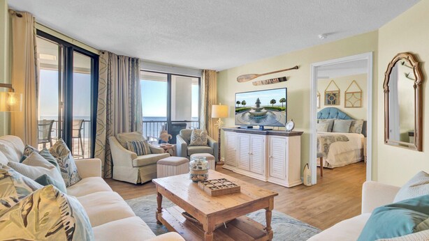 A spacious living room with a sofa, armchairs, a coffee table, and a television. There is a balcony with curtains, a dining area, and a bedroom with a bed and a nightstand.