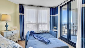 Une chambre à coucher comprenant un lit, une table de chevet, une lampe, une petite table et une vue sur la plage, visible à travers une grande fenêtre.