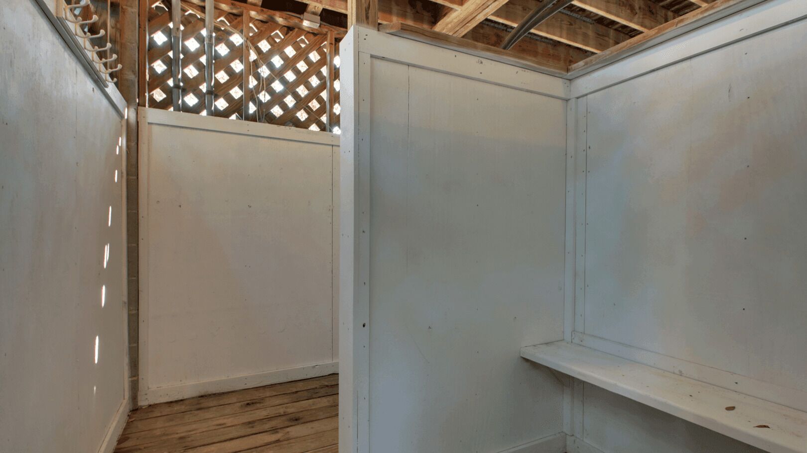 A room with white walls, a wooden floor, and a metal ceiling. There is a wooden lattice structure in the background.
