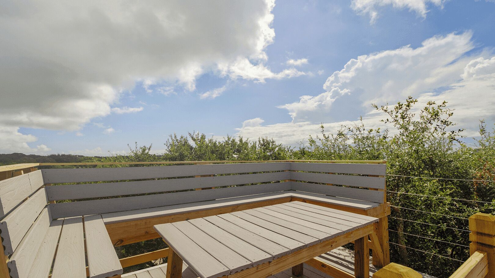 A wooden bench with a metal frame, set against a backdrop of a clear sky and greenery.
