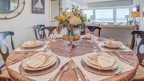 A round dining table set for six with a vase of flowers, a fruit basket, and a view of the ocean through large windows. - 1102 Ocean Club Avantstay Oceanfront Shared Pool, Spa Ocean Views (Isle of Palms)