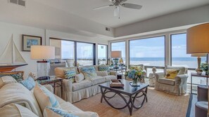 A spacious living room with a large window overlooking the ocean, a ceiling fan, and a cozy seating area with a coffee table. - 1102 Ocean Club Avantstay Oceanfront Shared Pool, Spa Ocean Views (Isle of Palms)
