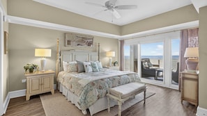 A bedroom with a large bed, a ceiling fan, a view of the beach through a sliding door, and a small balcony with a chair.