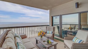 A balcony with wicker furniture, a table with a vase of flowers, and a bottle of champagne. - 1102 Ocean Club Avantstay Oceanfront Shared Pool, Spa Ocean Views (Isle of Palms)