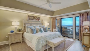 Une chambre à coucher comprenant un lit, des tables de chevet, un banc, un ventilateur de plafond et une vue sur l’extérieur, visible à travers une porte vitrée.