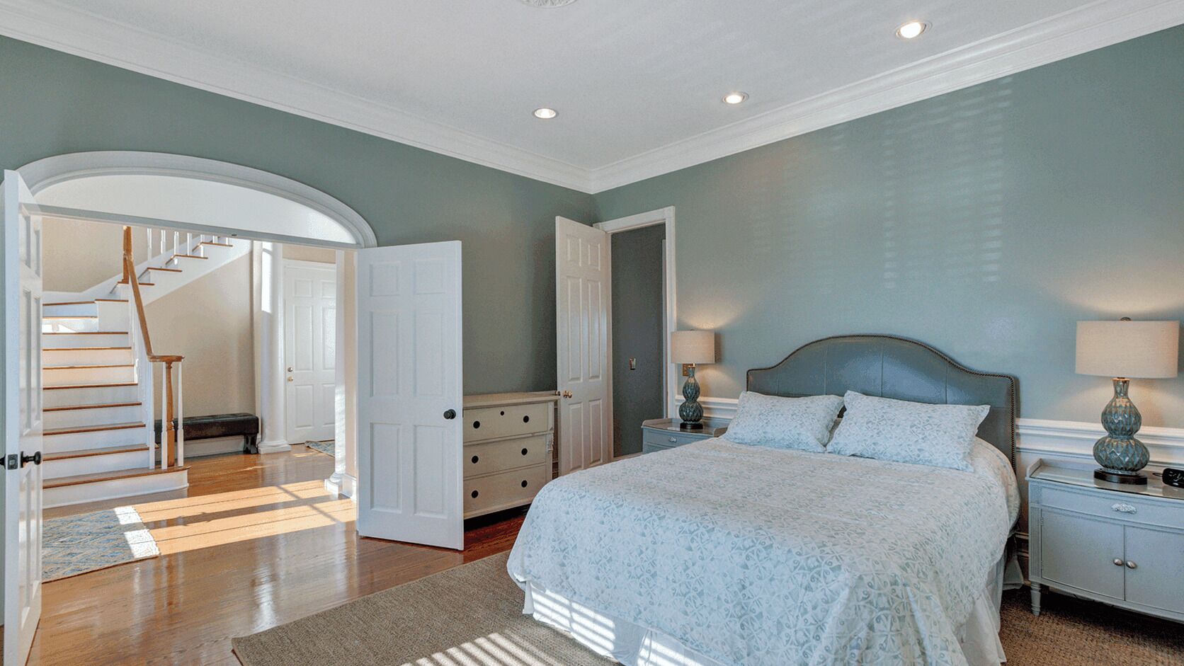 A bedroom with a bed, nightstands, a dresser, a staircase, and an open door leading to another room.
