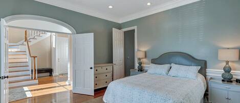 A bedroom with a bed, nightstands, a dresser, a staircase, and an open door leading to another room.