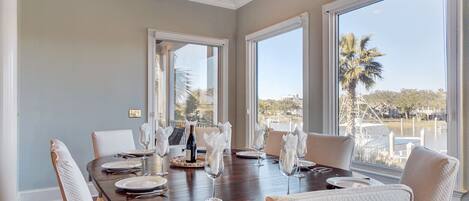 Un comedor con una mesa grande dispuesta para seis personas, ventanas amplias con vista a palmeras y un puerto deportivo, y una puerta que conduce a un área exterior.