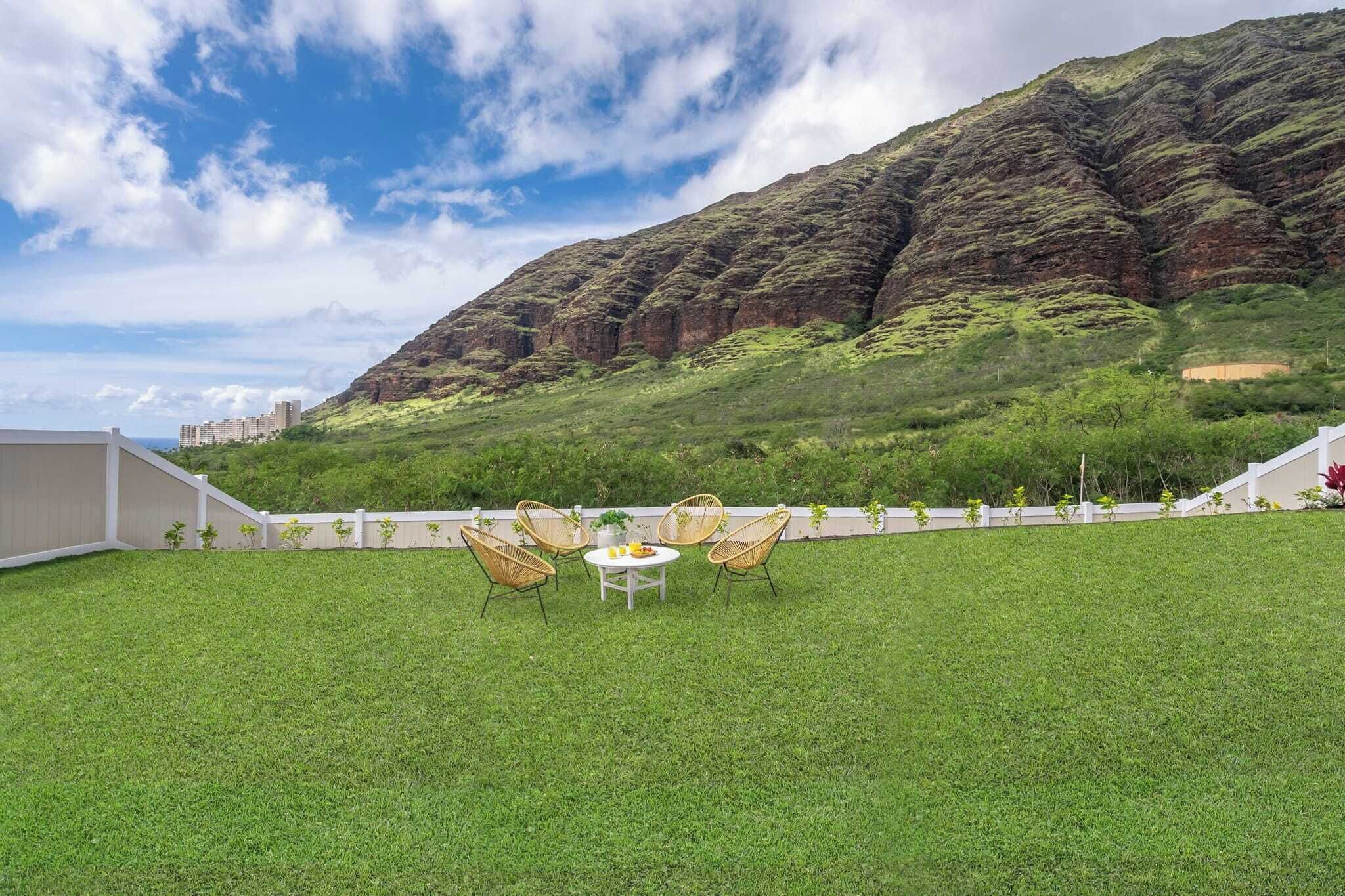A grassy outdoor area with a table and chairs, a white fence, and a mountainous backdrop.