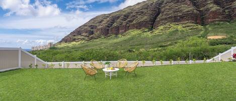 A grassy outdoor area with a table and chairs, a white fence, and a mountain in the background.