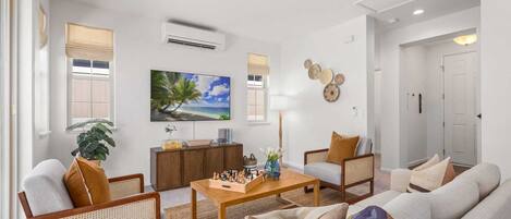 Una sala de estar con un sofá, sillas, una mesita de centro, un televisor y un cuadro con una playa.
