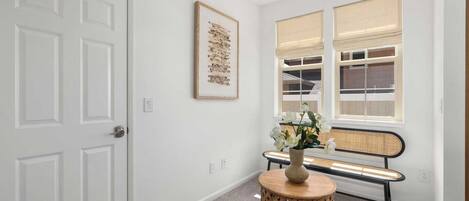 A hallway with a white door, a framed artwork on the wall, a bench with a woven seat, and a round table with a vase of flowers.