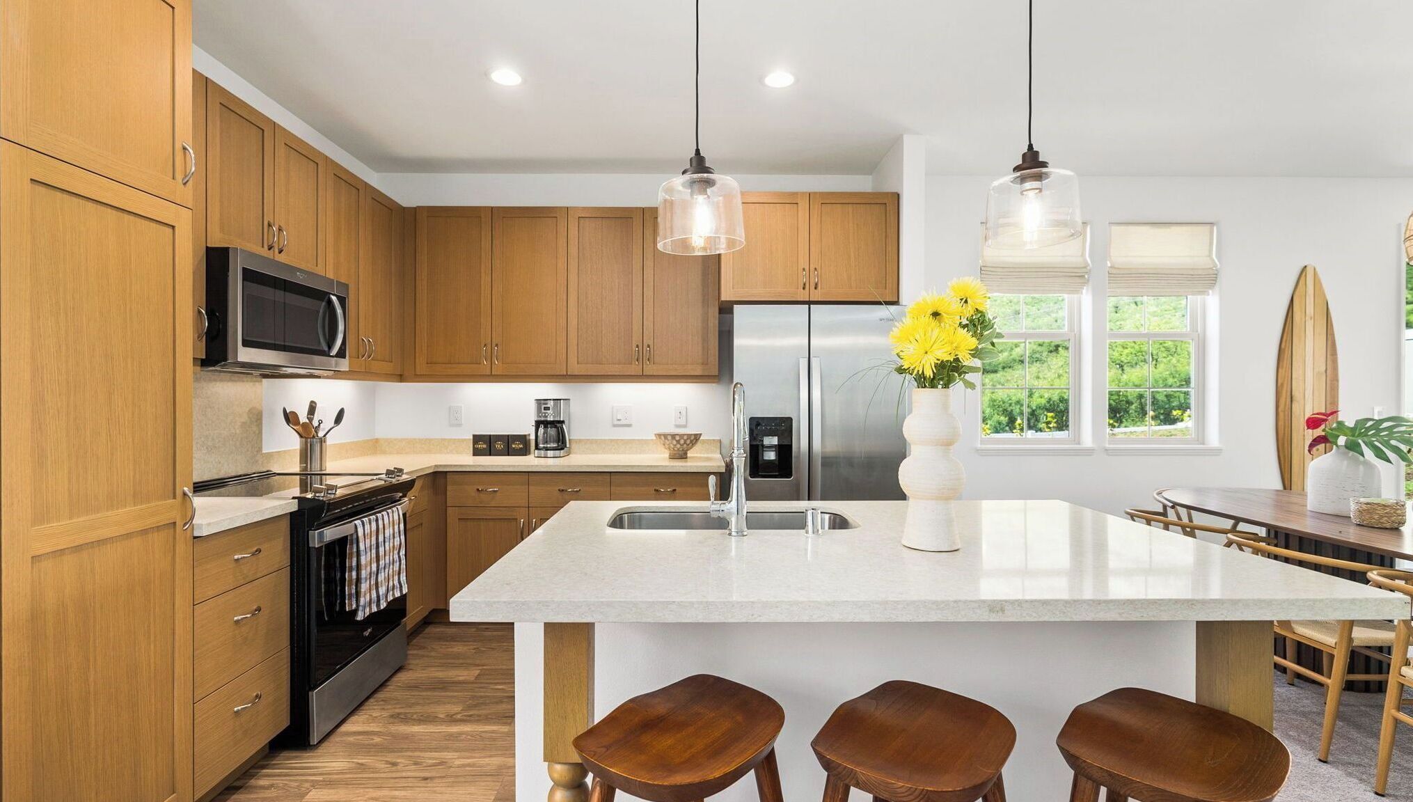 Une cuisine moderne avec des meubles en bois, des appareils en acier inoxydable, un îlot central doté de tabourets et une vue sur la verdure à l’extérieur.