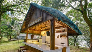 Un bar rústico al aire libre con techo de paja, taburetes de madera y vistas a un lago.