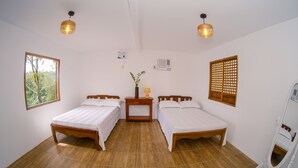 Una habitación con dos camas de madera, una mesita con lámpara, una ventana con vistas a árboles y un ventilador de techo.