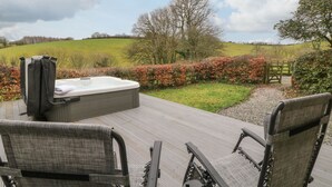 A hot tub on a wooden deck with chairs, surrounded by a landscaped garden and a view of the countryside. - The Hawthorns (South Molton)