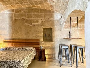 A rustic stone-walled room with a bed, a small shelf with bottles, and metal bar stools. - Casa Luisa (Monopoli)