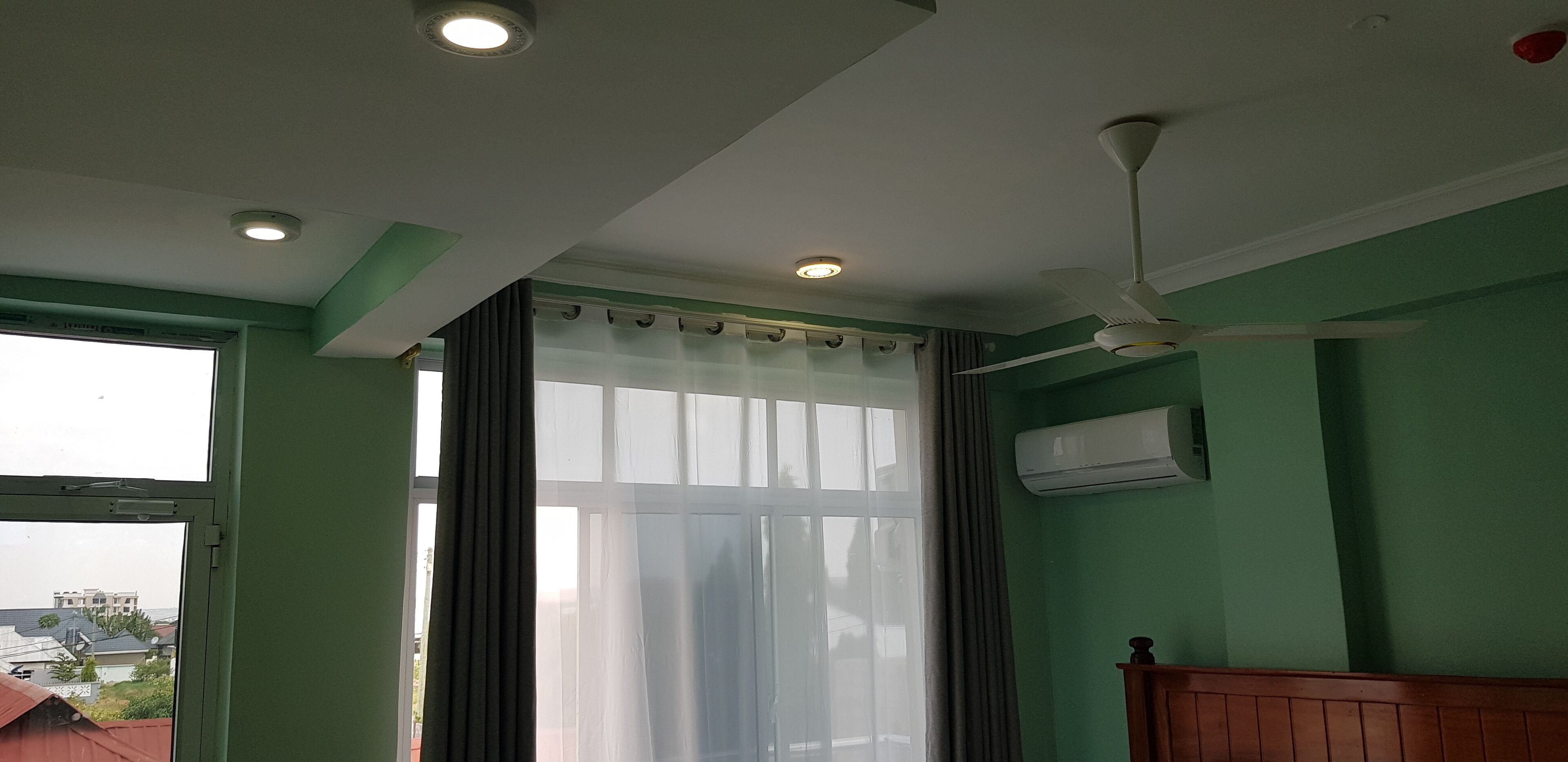 Una habitación con ventilador de techo, iluminación empotrada, una ventana con cortinas translúcidas y un equipo de aire acondicionado.
