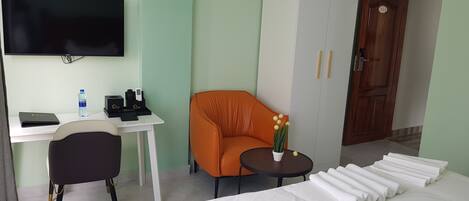 A hotel room with a bed, a desk with a computer, a chair, a small table with a plant, and a door with a number.