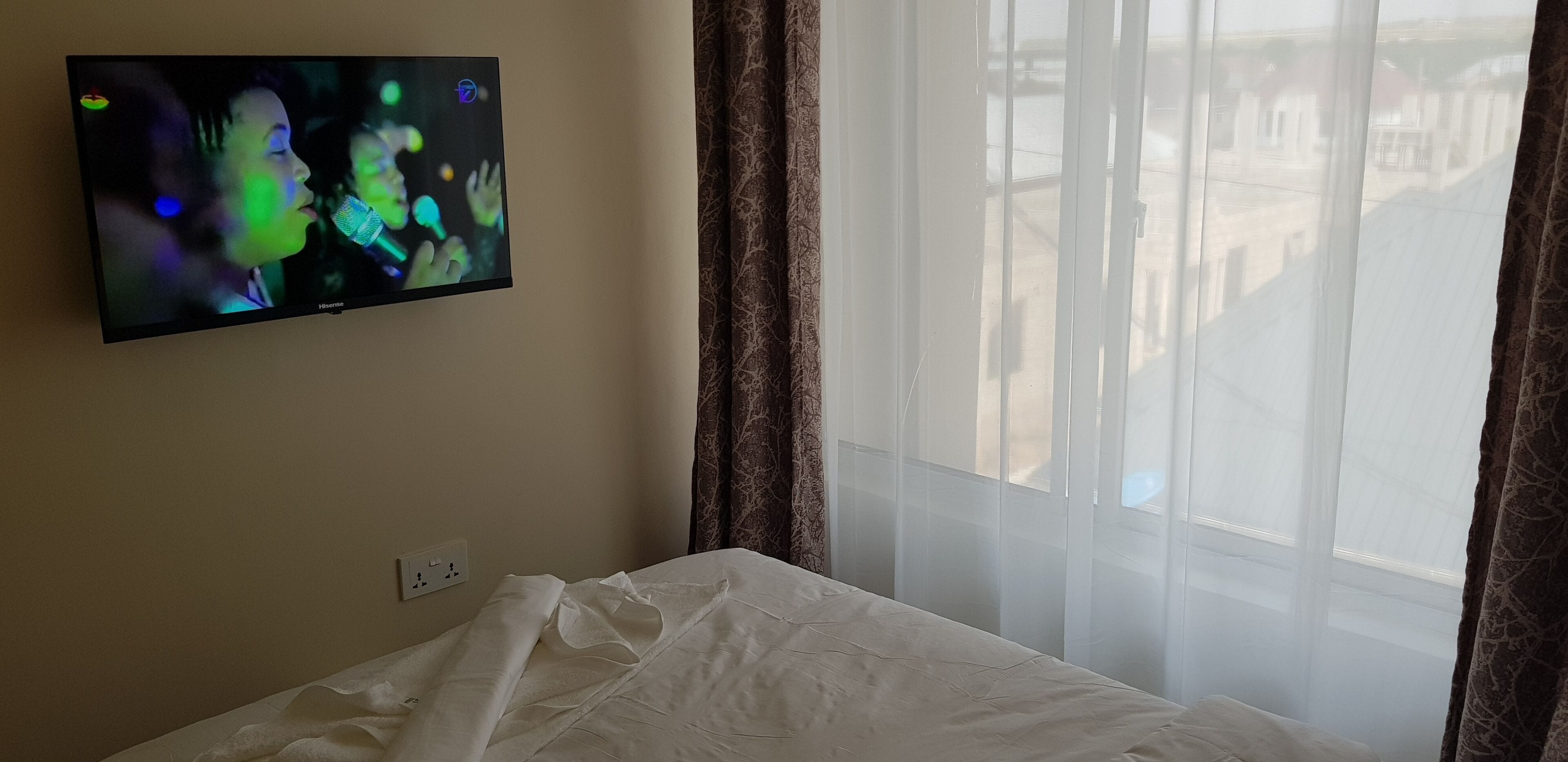 A hotel room with a bed, a television mounted on the wall, and a window with sheer curtains.