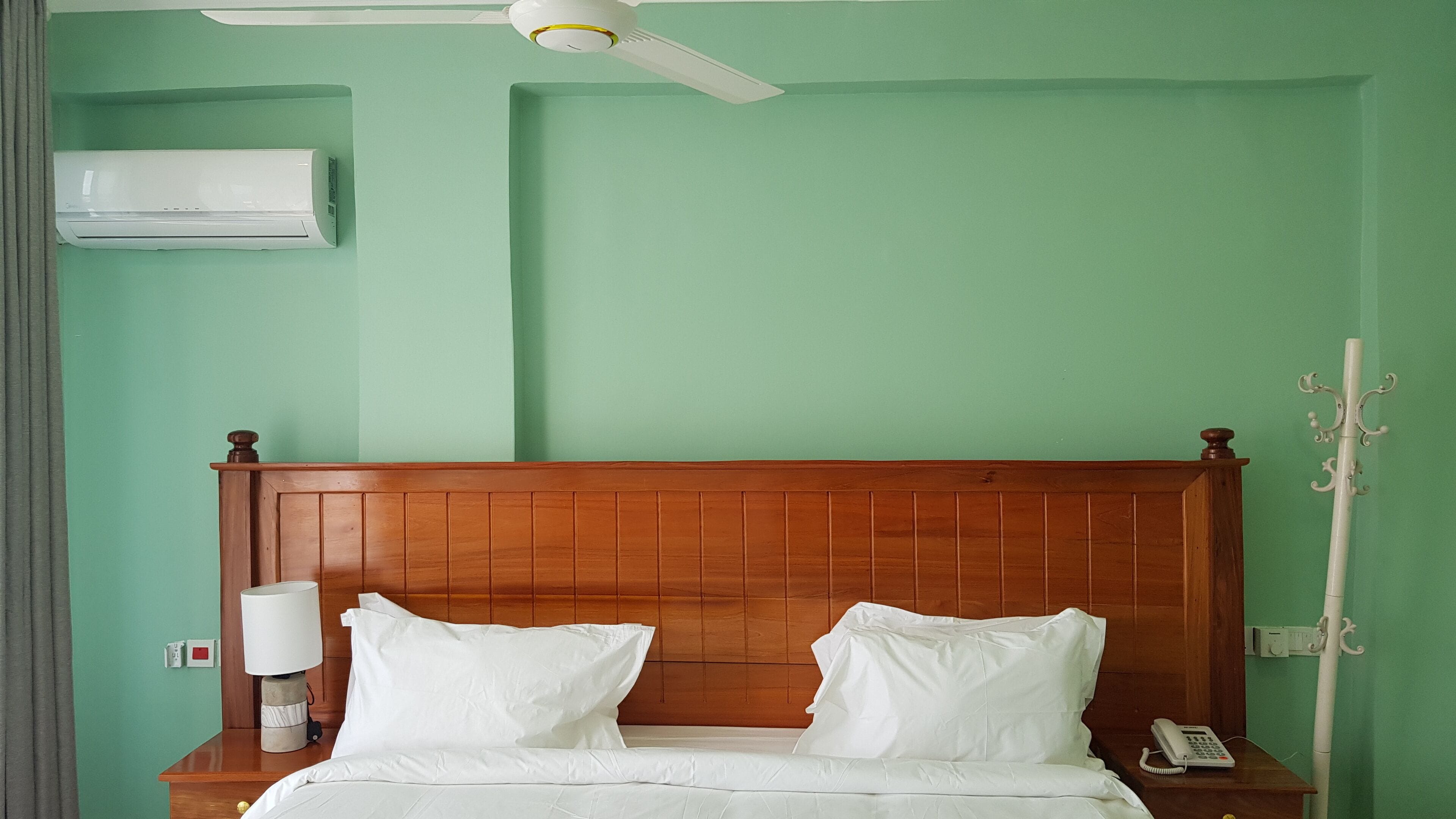 A hotel room with a wooden headboard, a bed with white linens, a bedside table with a lamp, a telephone, and an air conditioning unit on the wall.